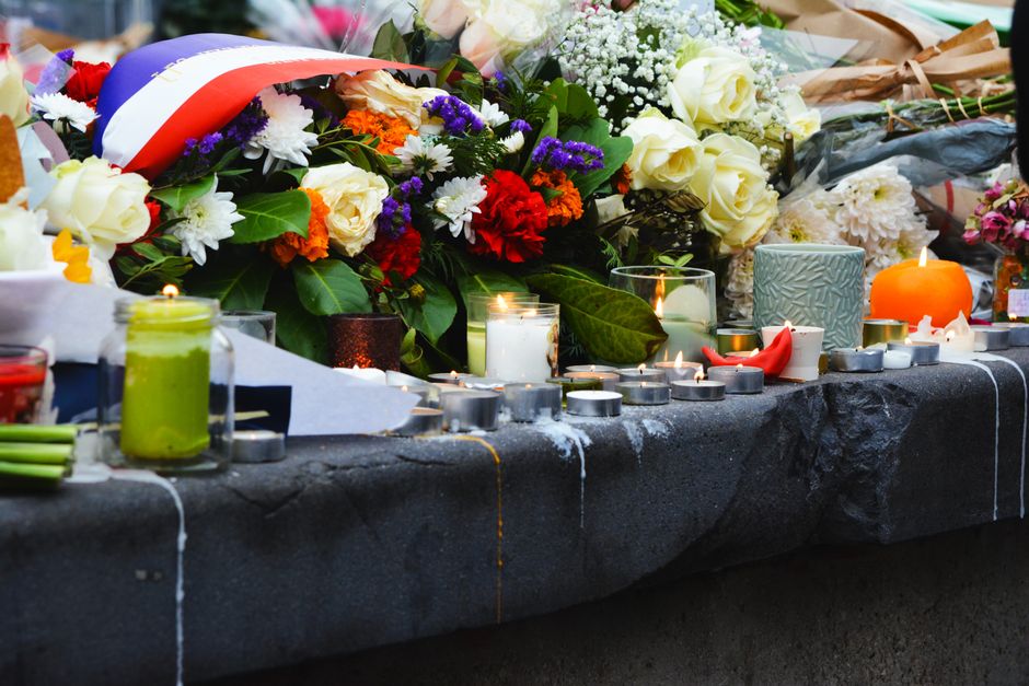 A floral tribute display on a dark surface, featuring a large bouquet of fresh flowers including white roses, red carnations, purple statice, and white daisies with green leaves. Several lit candles i