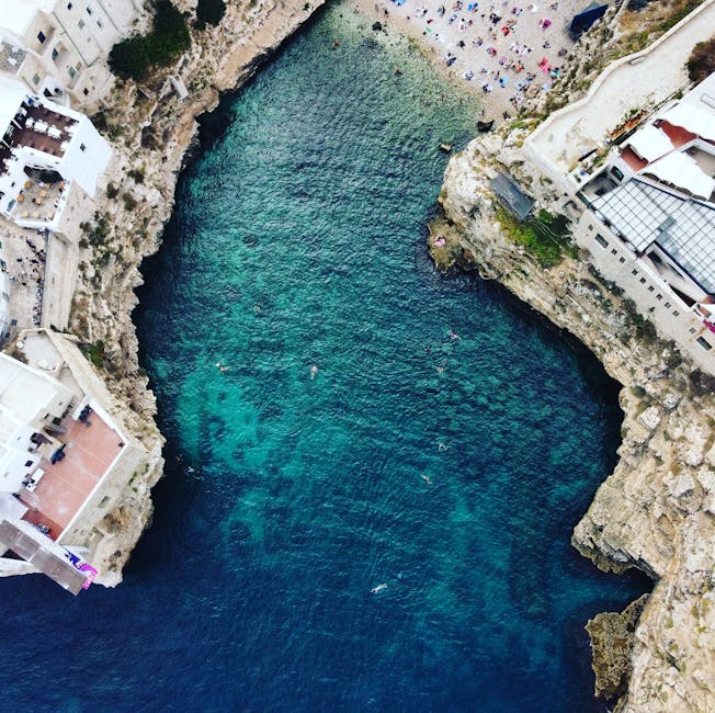Aerial view of a natural coastal inlet with clear, turquoise waters surrounded by white rocky cliffs. Several small boats are visible on the water's surface, with a few people swimming or snorkeling i