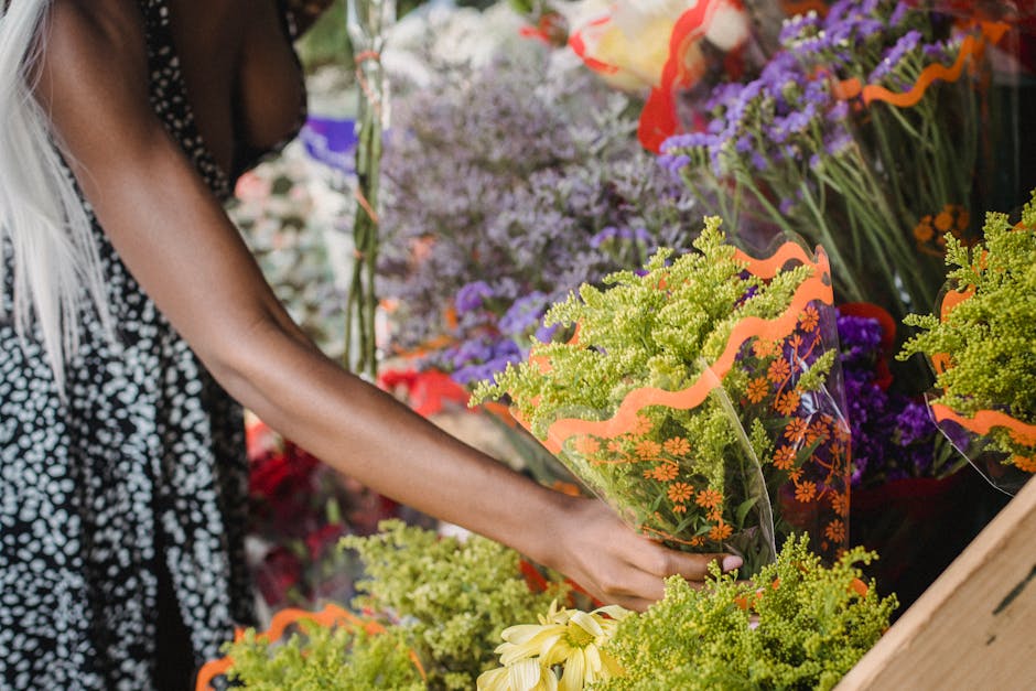 A person selecting a floral arrangement at a flower shop displaying a variety of fresh flowers, including yellow, purple, and red blooms. They are holding a bouquet wrapped in transparent cellophane w