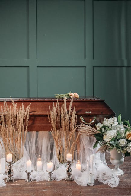 A floral arrangement featuring a bouquet of white roses and pale pink flowers resting atop a polished wooden casket, which is surrounded by cream-colored pampas grass and tall, clear glass candle hold