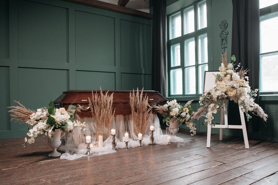 A floral arrangement featuring white roses, lilies, and other delicate white blooms, complemented by soft beige pampas grass and lush green leaves, arranged in two white ceramic vases and placed on ei
