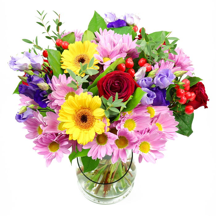 In this image, a vibrant mixed bouquet overflows from a clear glass vase, set against a bright white background that makes every petal, leaf, and stem easy to distinguish. The composition is generous and rounded, with deep red roses forming the central focus; their layered, velvety petals unfurl with a sense of classic romance and quiet strength. Flanking the roses are striking yellow gerbera daisies, each bloom with a dark brown-orange centre ringed by golden petals that stretch outward like rays of warm sunlight. Soft pink daisies or chrysanthemums drift around these central flowers, their many fine petals and petite yellow centres creating a cushion of colour that feels light and uplifting. Threaded through the pinks are stems of lisianthus in shades of purple and lavender, their bell-shaped flowers and partially opened buds introducing a cool, sophisticated tone that adds depth and dimension to the bouquet. Scattered among the petals are bright red hypericum berries, small and rounded with a glossy sheen that catches the light, adding both texture and a playful, almost jewel-like detail. The entire design is woven together with varied greenery: broad, darker green leaves that form a sturdy base, and more delicate, lighter sprigs that curl and soften the edges, echoing the wild yet gentle greenery you might notice around Hackney Marshes on a calm afternoon. Through the transparent vase, cut stems are visible in clear water, neatly aligned to show the care and technique behind the arrangement. This bouquet feels joyful and thoughtful, ideal for birthdays, thank-you gestures, or simply bringing a burst of fresh colour into a Hackney Marshes living room or workspace.